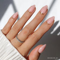 A hand with short almond nails in glamnetic’s Goal Chaser, a light pink polish with minimalist gold half-moon and French tip designs. Two thin, stacked rings with small gems are worn. The white ribbed sleeve completes the softly lit look.