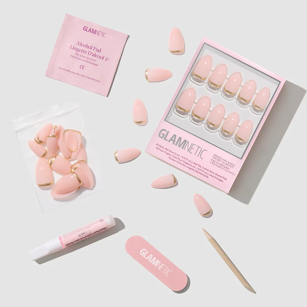 Flat lay of the glamnetic Goal Chaser pink press-on nail kit, with short almond and gold French tip nails, plus a nail file, cuticle stick, glue, alcohol pad, and extra nails on a light gray background.