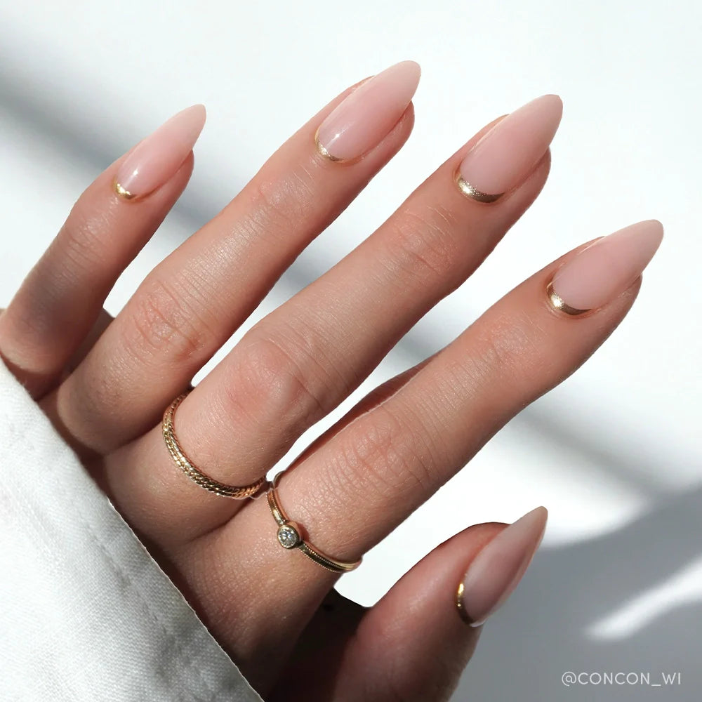A hand with medium almond nails in glamnetic's Goal Getter—light nude polish with thin gold French tips—wears two gold rings against a softly lit white background.