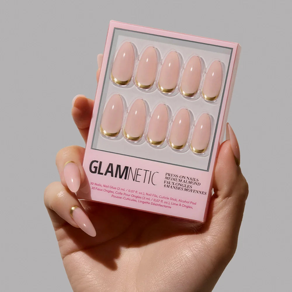 A hand with medium almond nude pink nails holds a box of glamnetic Goal Getter press-on nails, featuring elegant gold French tips, against a plain gray background.