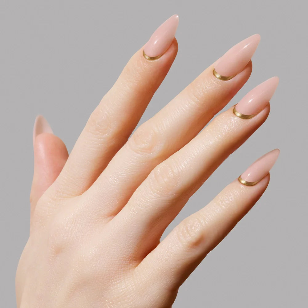 A hand wears glamnetic’s Goal Getter medium almond nude press-on nails, each with a minimalist gold accent near the cuticle, displayed against a plain light gray background.
