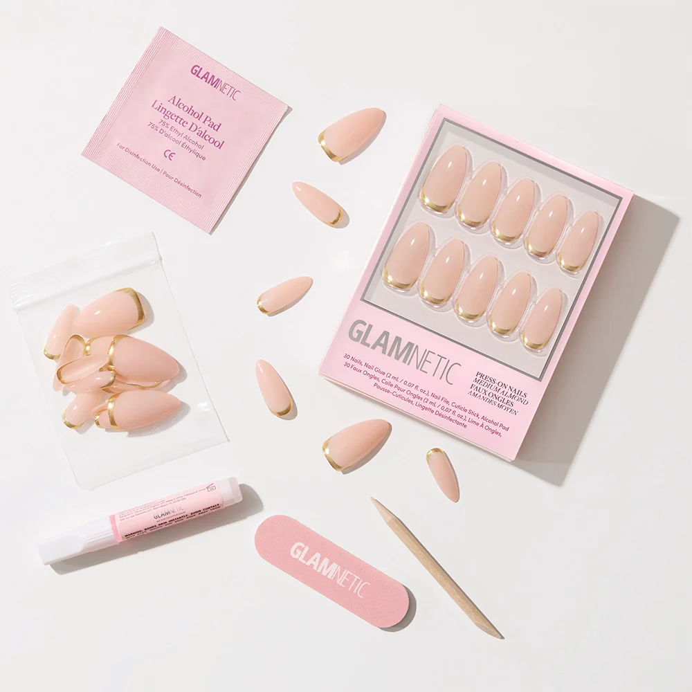 The glamnetic Goal Getter press-on nail kit includes nude almond-shaped nails with gold French tips, adhesive, a pink nail file, cuticle stick, alcohol pad, and spare nails—all arranged neatly on a white background.