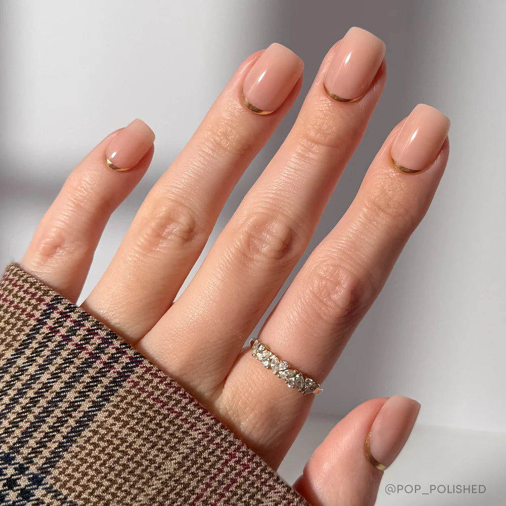 A hand with short squoval nude nails in "Goal Setter" by glamnetic, featuring a thin gold base accent like gold French tips. The person wears a plaid sleeve and a gold ring with clear stones on the ring finger against a softly lit background.