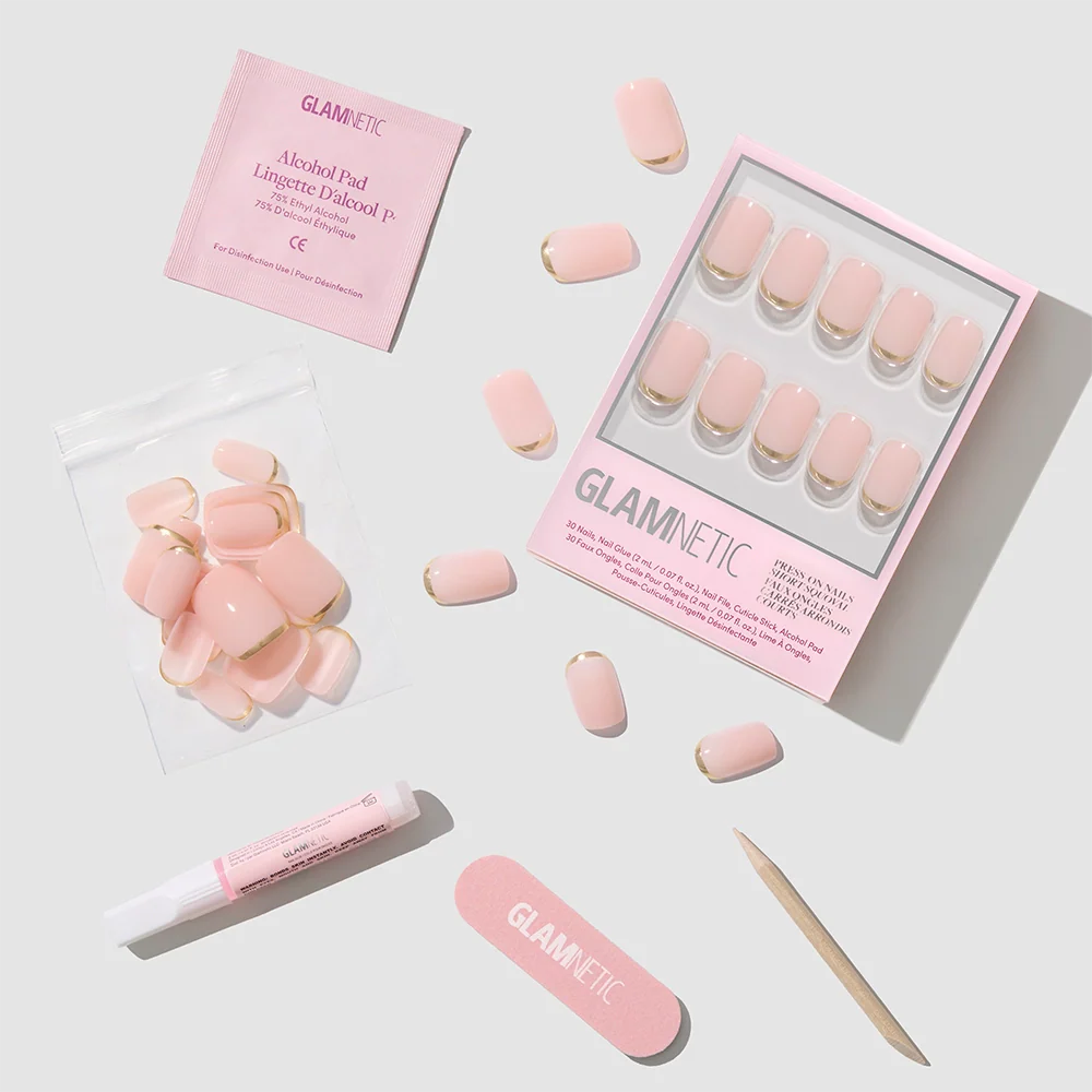 The Goal Setter press-on nails by glamnetic feature short squoval shapes in light pink with gold French tips. Included are an alcohol pad, nail glue tube, wooden cuticle stick, pink nail file, and extra nails—displayed on a light gray background.