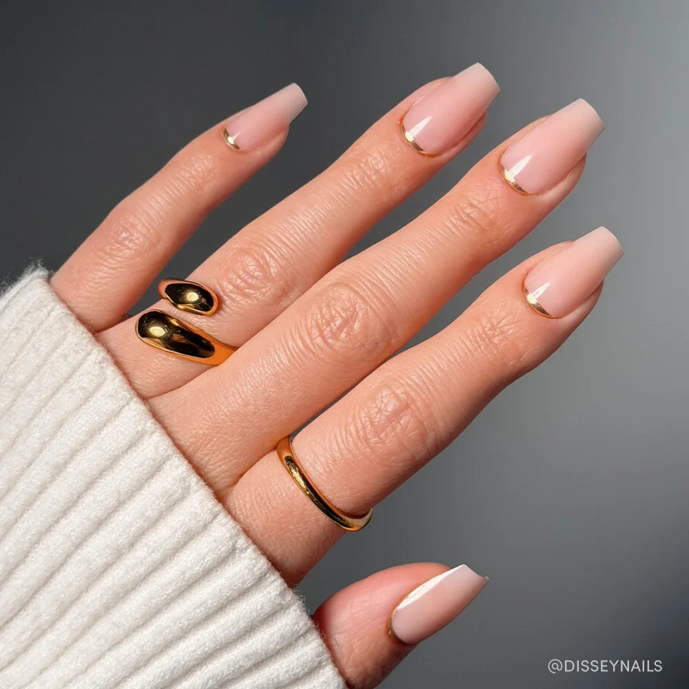 A hand with square-shaped nude pink Gold Goals Press-On Nails by glamnetic, featuring thin gold crescent accents near the cuticles, is accessorized with three gold rings and a cream ribbed sleeve for a chic salon-quality look.