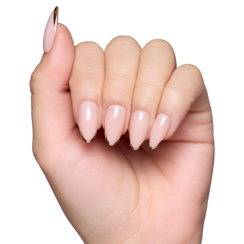 A hand with long, almond nails in glamnetic’s Gold Standard—a glossy light pink set with thin metallic gold tips—shines against a white background.