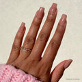 A hand with glamnetic’s “In The Nude” square-shaped, nude-pink ombré nails appears against a light background. Two gold rings adorn the fingers, while a pink knitted sleeve covers the wrist. This vegan, cruelty-free manicure is minimalist.