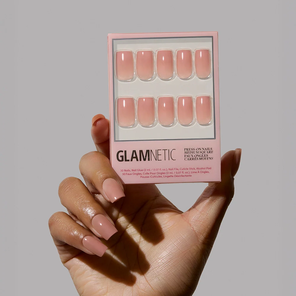 A hand with long, glossy, nude-pink ombré nails holds a box of glamnetic In The Nude press-on nails against a plain light gray background, highlighting the perfect minimalist manicure.