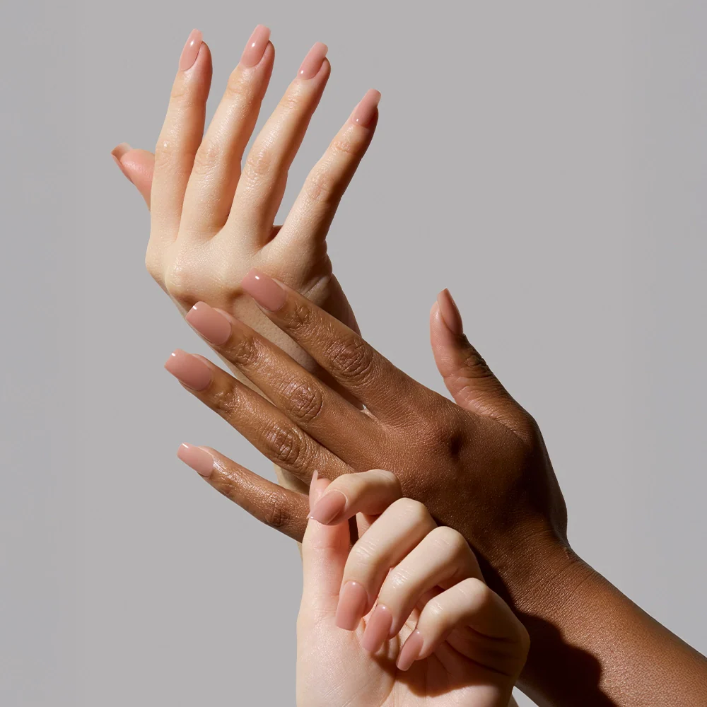 Three hands with varying skin tones display neatly manicured nails painted in glamnetics In The Nude, a nude-pink ombré polish, gracefully posed together against a plain light gray background.