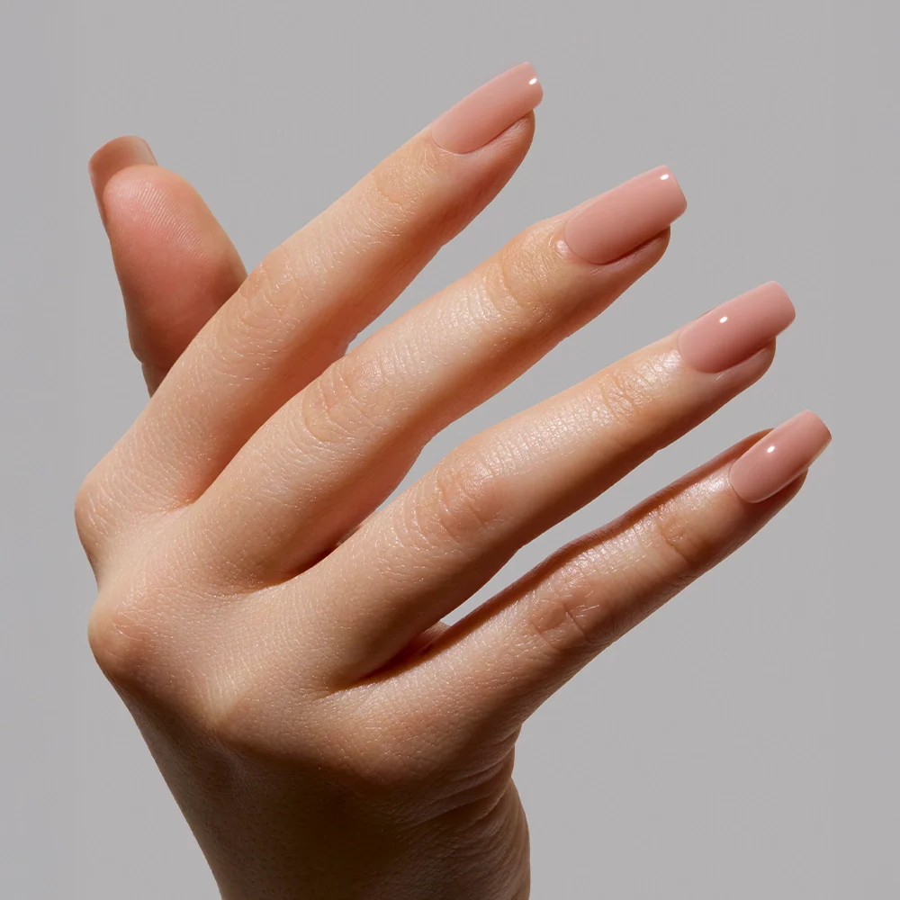A close-up of a hand with long, square nails in a glossy nude-pink ombré minimalist manicure, featuring In The Nude by glamnetic, against a plain light gray background.