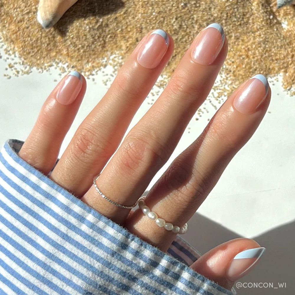 A hand with glamnetic’s Into Blue salon-quality periwinkle micro French tips rests near sand and a seashell. The person wears a striped shirt and two delicate rings, one with pearls and one plain.