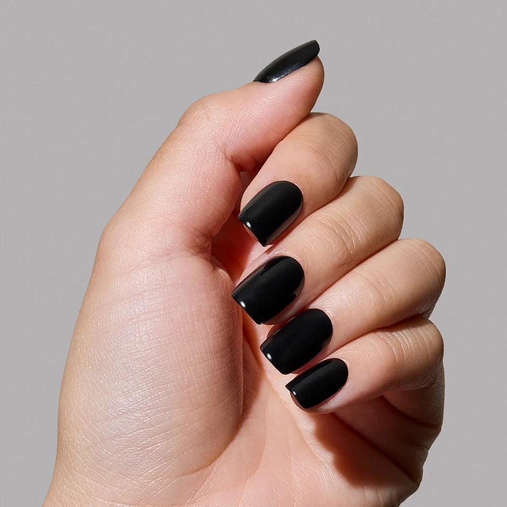 A hand with glossy squoval nails painted in glamnetics Jet Black is held against a plain light gray background.