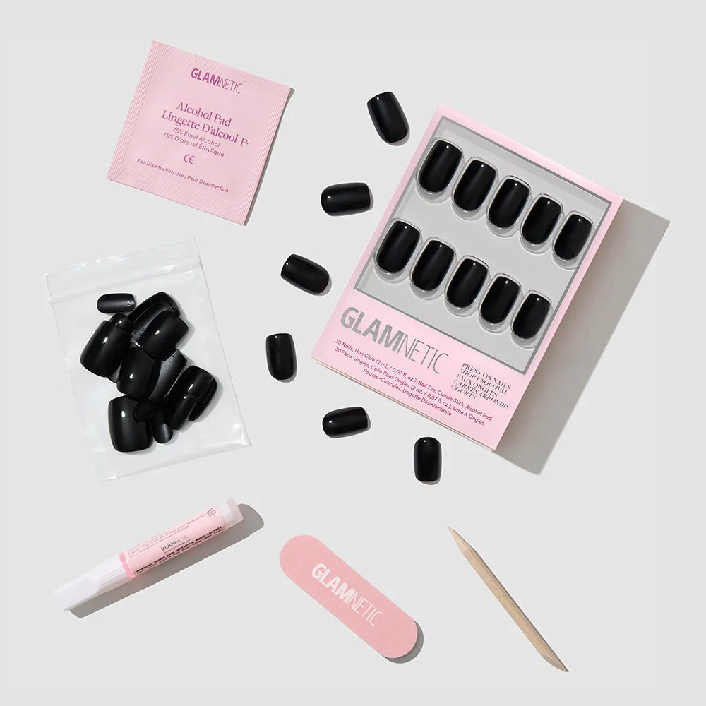 A flat lay of the glamnetic Jet Black set displays glossy squoval nails, nail glue, a wooden cuticle stick, pink file, alcohol pad, and clear bag—all you need for reusable vegan nails—on a light gray background.