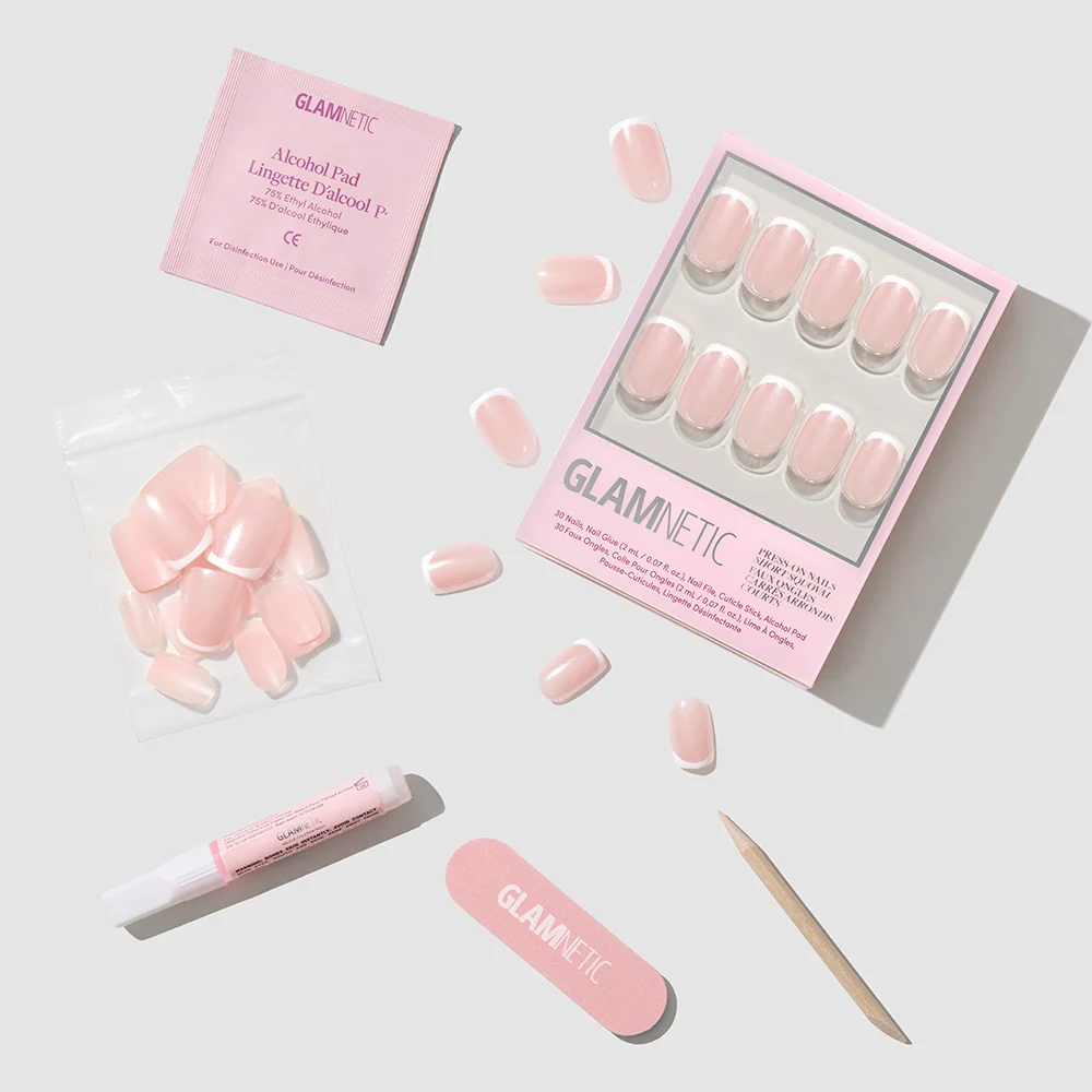 A flat lay showcases the glamnetic La Perle Nail Set with short squoval French tip nails, extra nails, a pink nail file, cuticle stick, glue tube, and alcohol pad arranged on a light gray background.