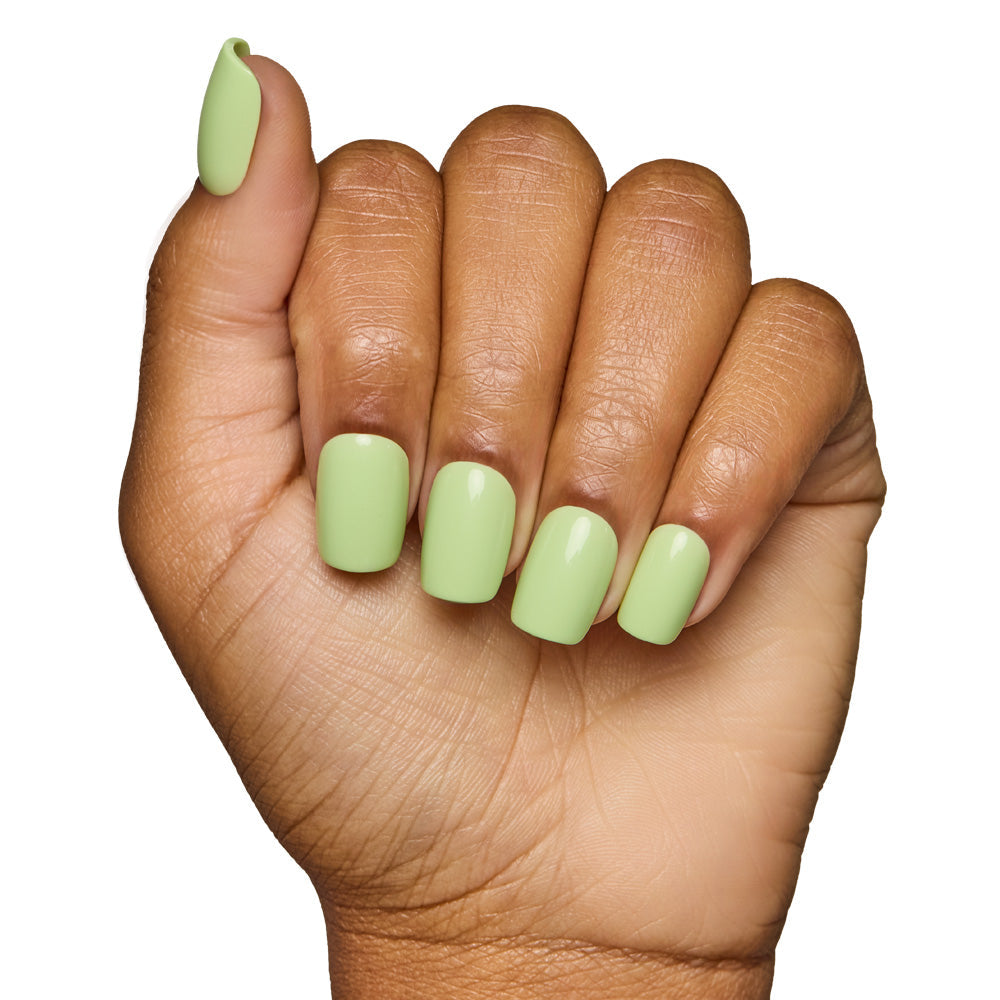 A hand with medium brown skin shows fingers curled inward, featuring neatly manicured short squoval nails painted in glamnetics Light Sage, a glossy pale green shade. The background is plain white.