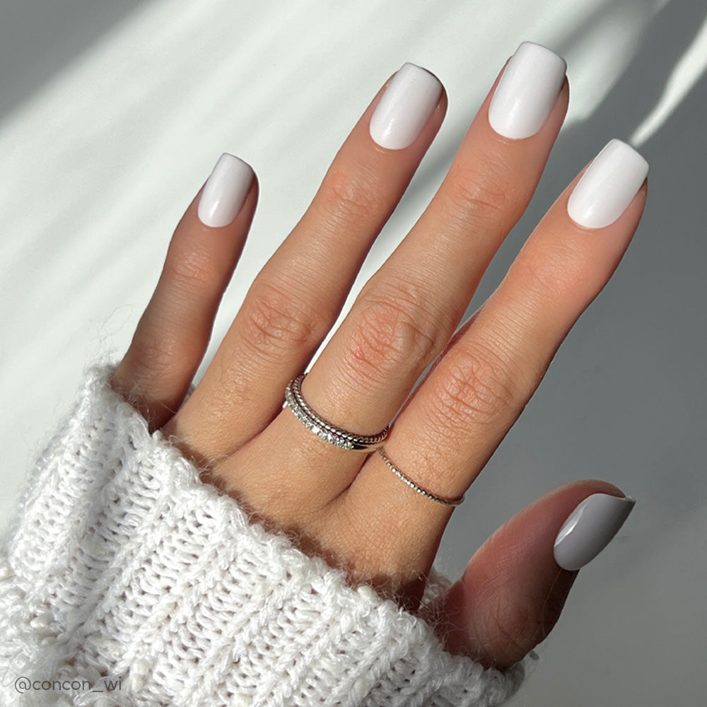 A hand in a white sweater sleeve displays glossy Lilac Gray squoval nails by glamnetic, complemented by two thin silver rings on the middle and ring fingers. The soft lighting adds elegance to the overall look.