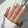 A hand in a white sweater sleeve displays glossy Lilac Gray squoval nails by glamnetic, complemented by two thin silver rings on the middle and ring fingers. The soft lighting adds elegance to the overall look.