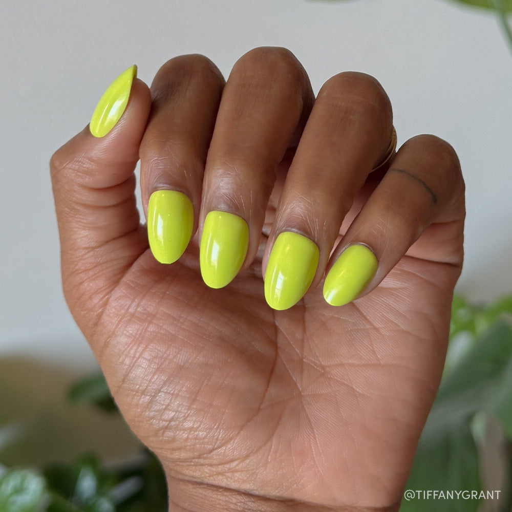 A hand with medium brown skin displays the glamnetic Lime Green short oval nails in a bold neon yellow-green shade, set against a blurred green foliage background.