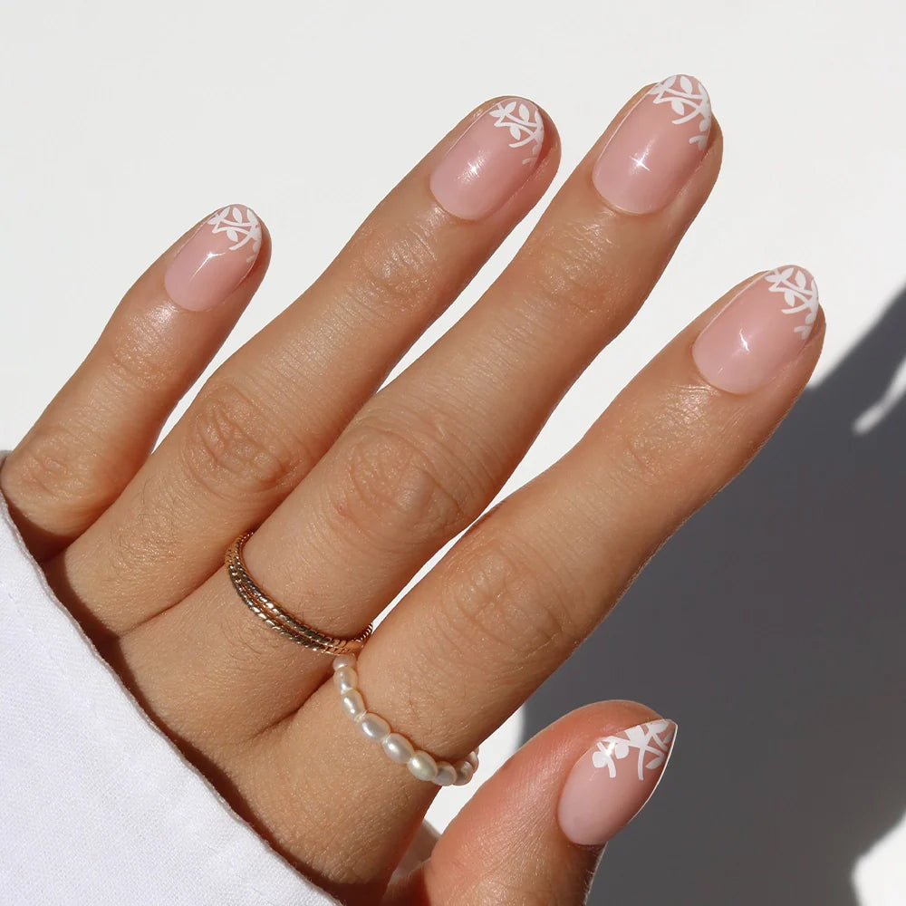 A hand with manicured nails features glamnetic's Love Preserved—natural pink press-ons with white leaf tip designs. The person wears gold and pearl rings and a white long-sleeve top.