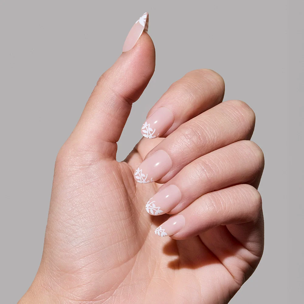 A hand with manicured nails in glamnetic’s Love Preserved style—nude base and delicate white leaf tip design—shown against a plain gray background.