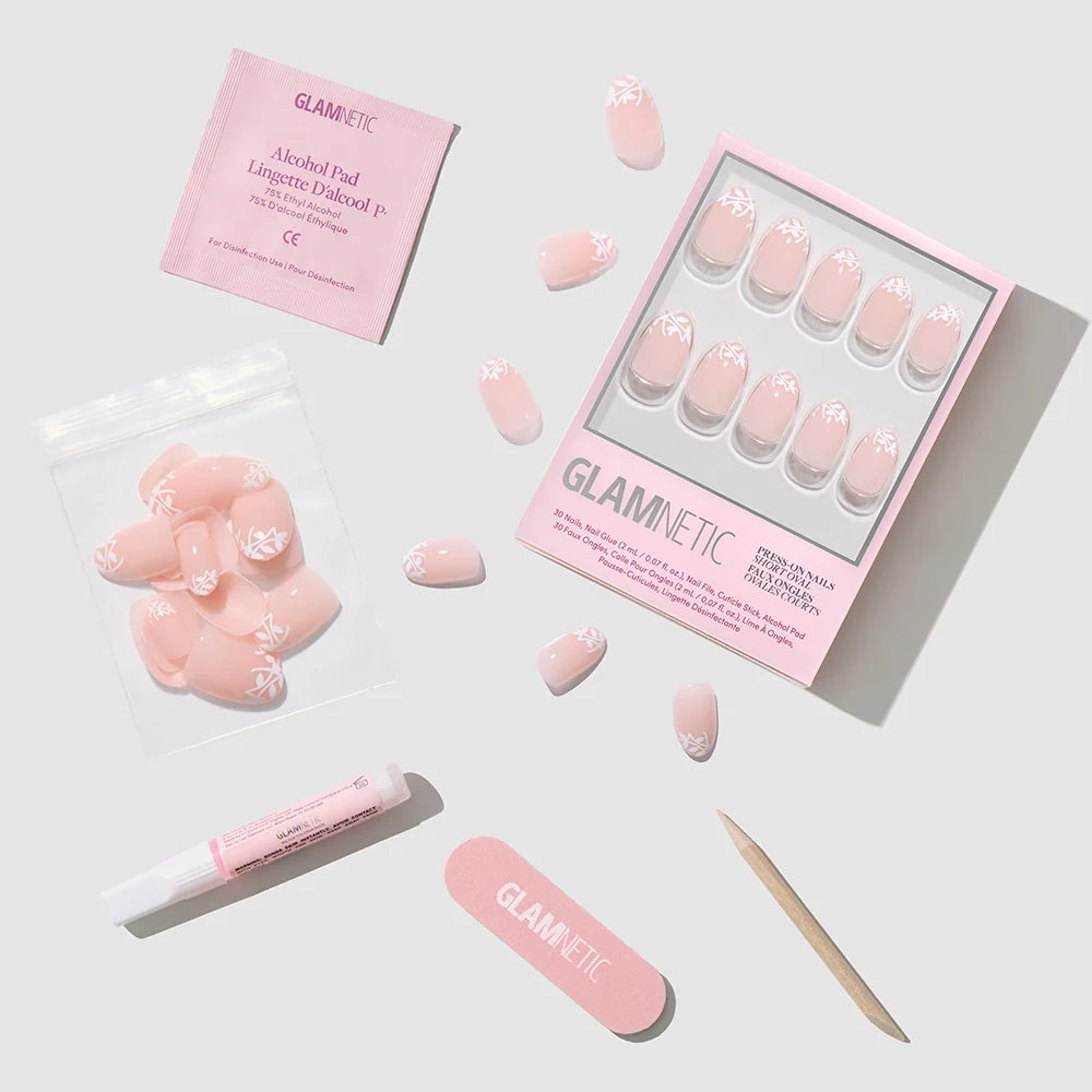 A flat lay of the Glamnetic Love Preserved press-on nail set shows pink botanical nails with a white leaf design, a nail file, cuticle stick, nail glue, alcohol pad, and extra nails in a bag on a light gray surface.