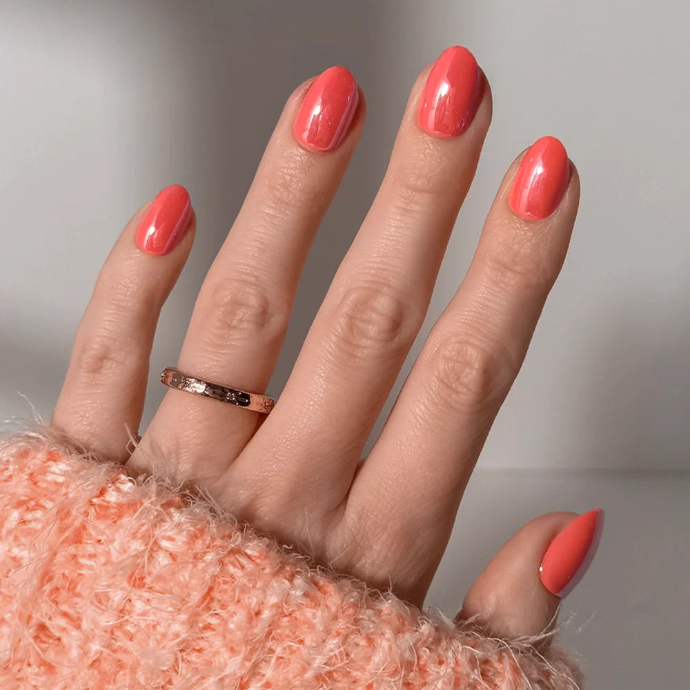 A hand with glamnetic’s Lush Coral glazed short oval nails, adorned with a gold ring and a soft peach fuzzy sweater, is pictured against a simple light background.