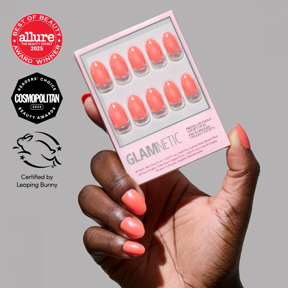 A hand with vibrant coral nails holds a box of glamnetic Lush Coral vegan, cruelty-free press-ons. Allure, Cosmopolitan, and Leaping Bunny certification logos are visible.