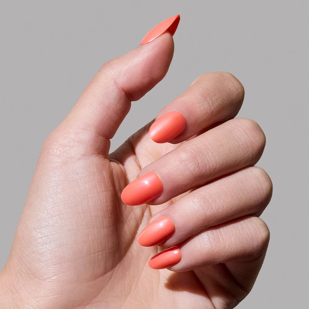 A hand with medium-length oval nails painted in glamnetic’s Lush Coral, a smooth, glossy vibrant coral polish, against a plain gray background.