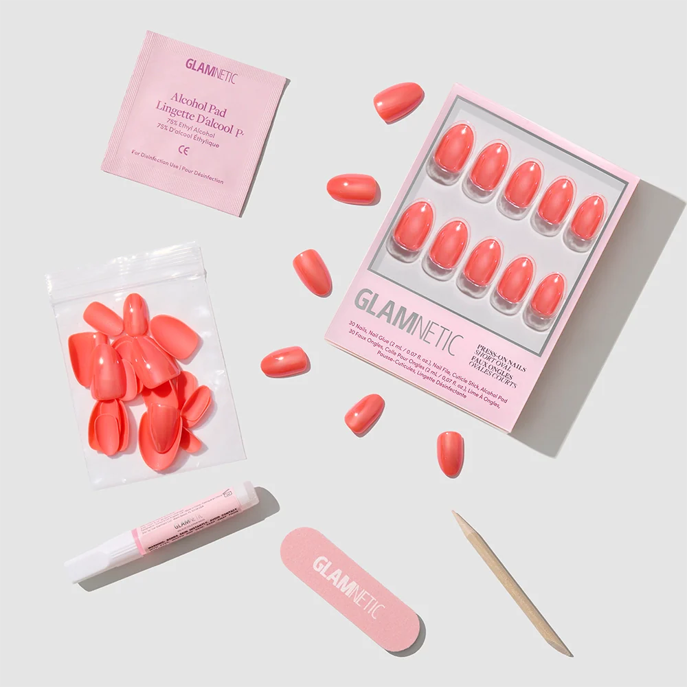 The Lush Coral set by glamnetic includes vibrant coral short oval nails, extra nails, nail glue, a wooden cuticle stick, an alcohol pad, and a nail file—all arranged neatly on a light surface with the glamnetic logo visible.