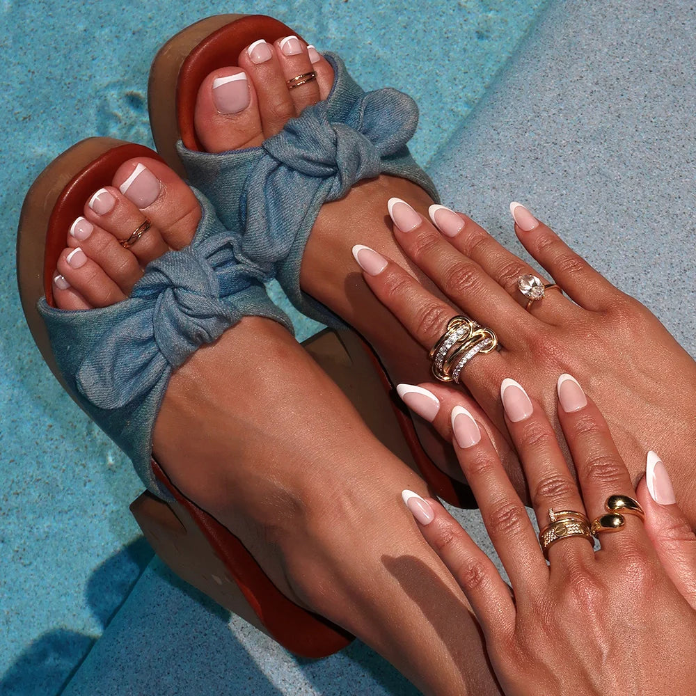 Feet and hands with matching white French-tip Glamnetic nails, worn with stacked gold rings and soft denim wedge sandals.