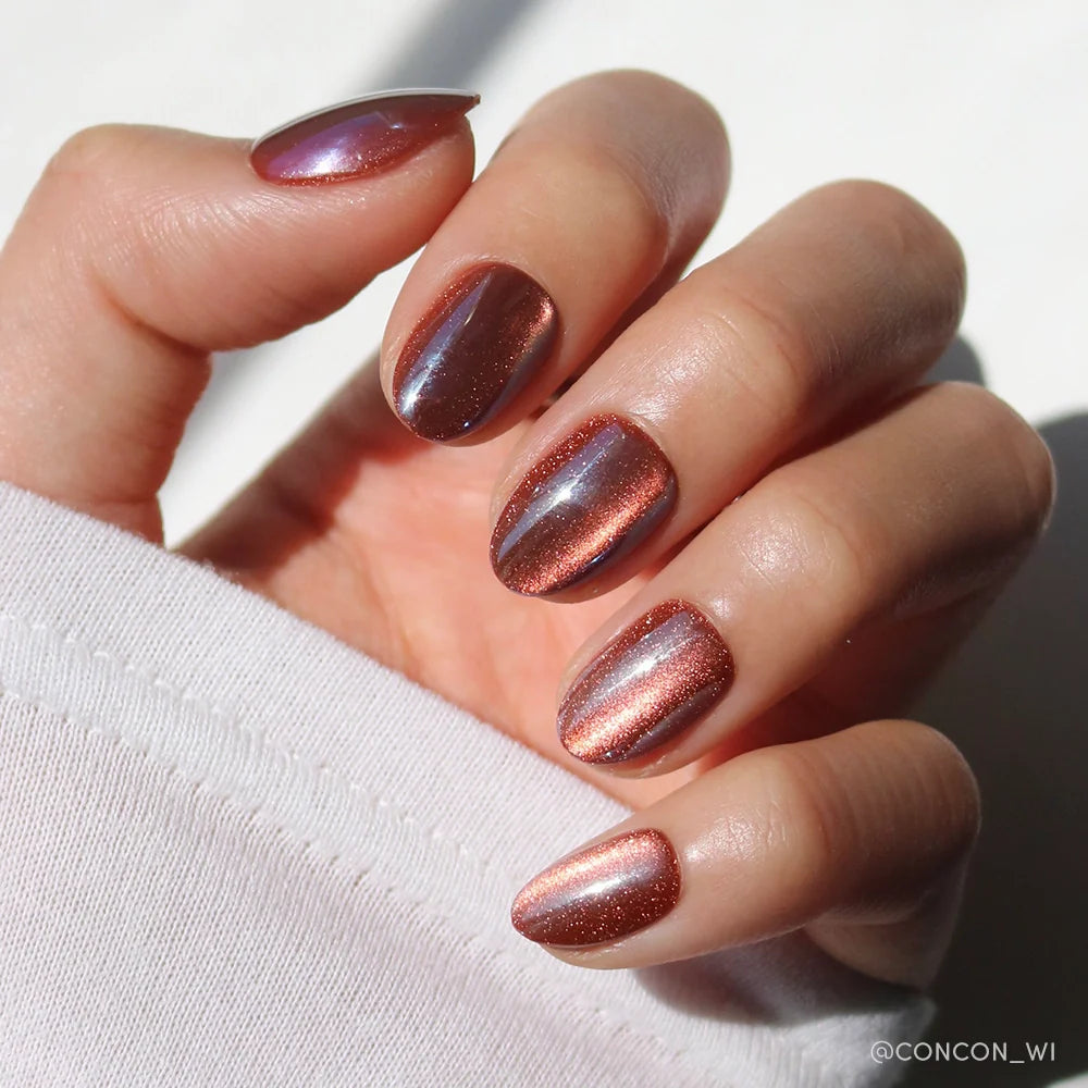 A hand with short oval nails painted in glamnetic’s Magnetic Moscow, a shiny metallic rose gold, glows in sunlight. The person wears a white long-sleeve shirt, highlighting the sets chic elegance.