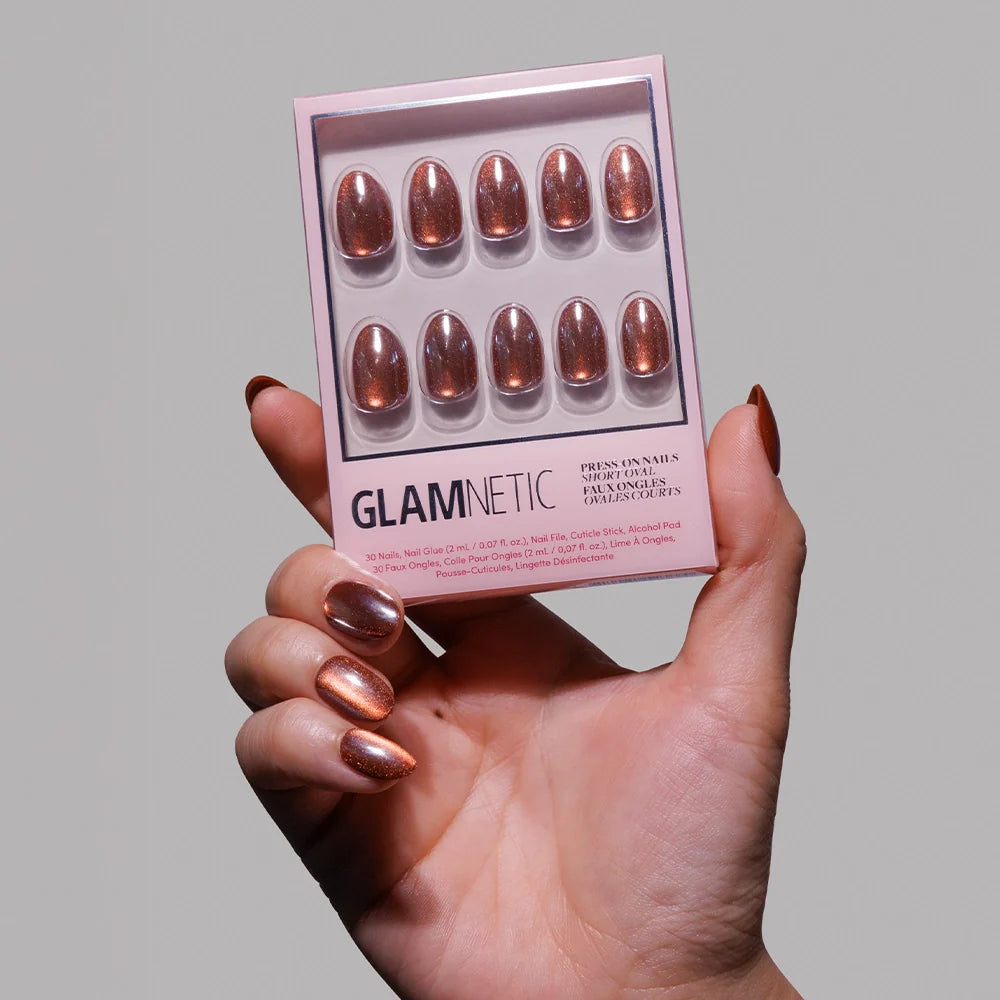 A hand with shiny, metallic copper short oval nails holds a glamnetic Magnetic Moscow nail set in a matching shade against a plain gray background.