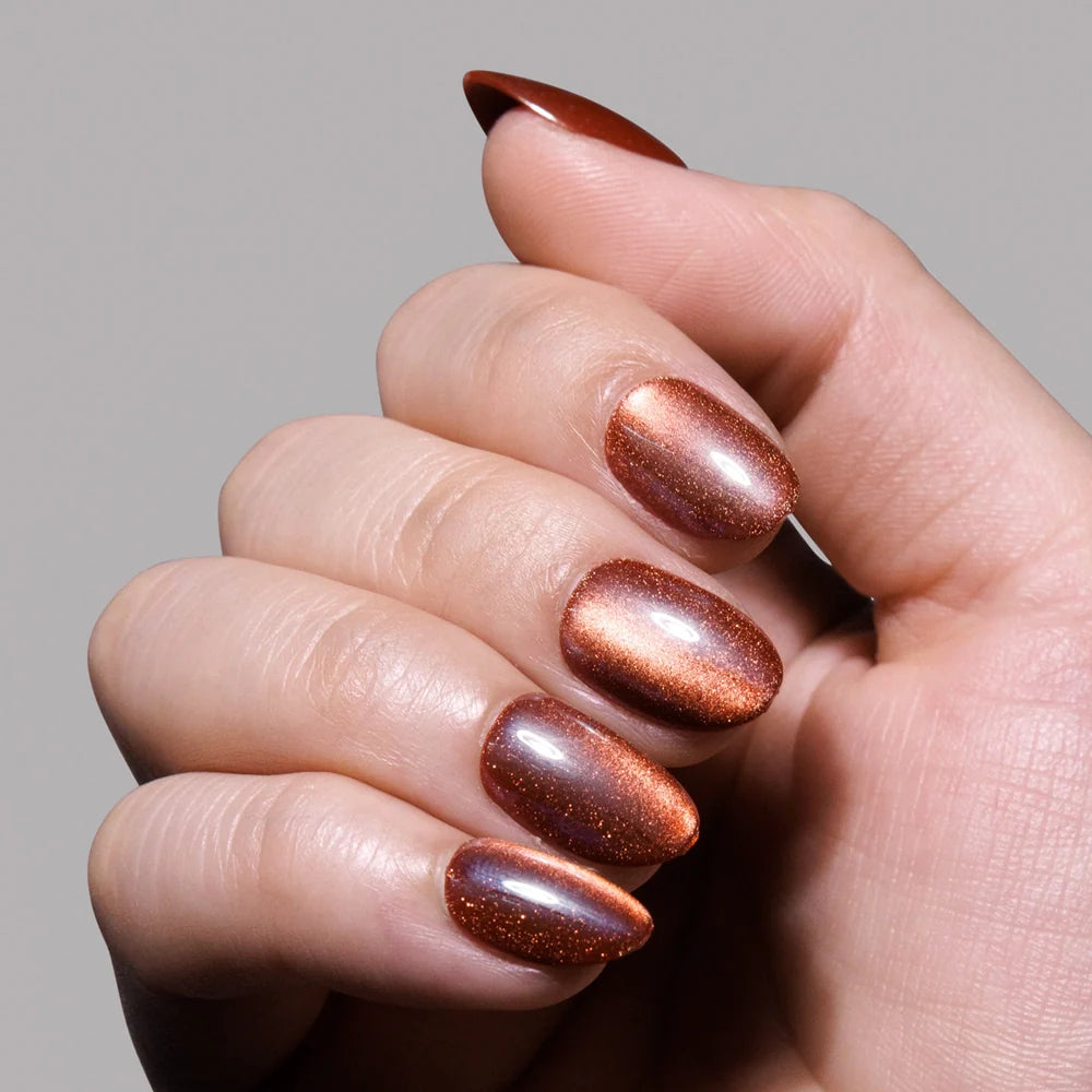 A hand with almond-shaped cat-eye nails painted in glamnetic’s Magnetic Moscow, a shimmery metallic copper shade, shown against a plain gray background.