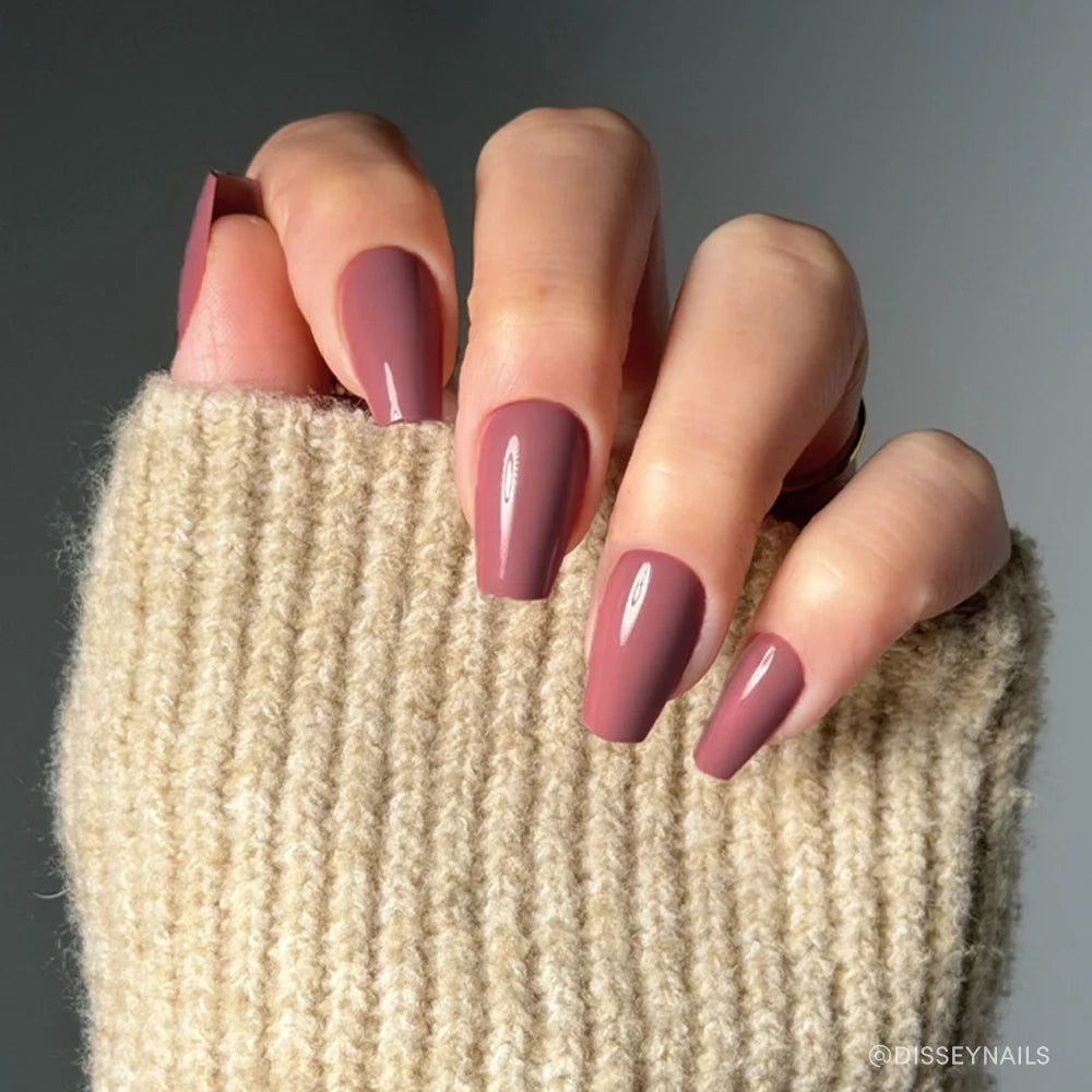 A hand with long, medium coffin nails painted in glamnetic’s Mauve Affair shade, partially covered by the sleeve of a chunky beige knit sweater.