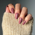 A hand with long, medium coffin nails painted in glamnetic’s Mauve Affair shade, partially covered by the sleeve of a chunky beige knit sweater.