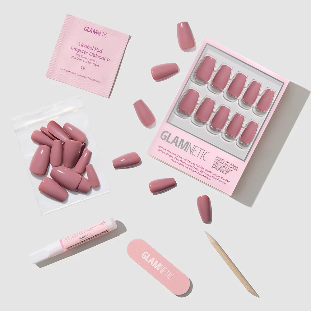 Mauve Affair by glamnetic: glossy pink medium coffin fake nails, nail file, wooden cuticle stick, glue tube, alcohol pad, and extra nails—all arranged on a light gray surface with glamnetic packaging.