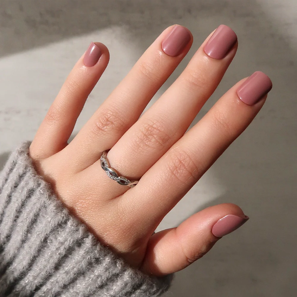 A hand with super short, square-shaped nails painted in glamnetic's Mauve Cutie polish, wearing a silver ring with small stones and a gray ribbed sweater, is shown against a light background.