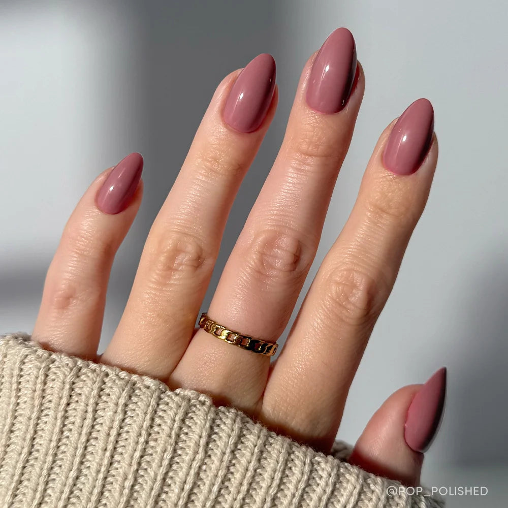 A hand with neatly manicured, short almond nails painted in glamnetic's Mauve Obsession—a dusty rose polish with a glossy finish—wears a gold chain ring and is partially covered by a beige knit sweater sleeve.