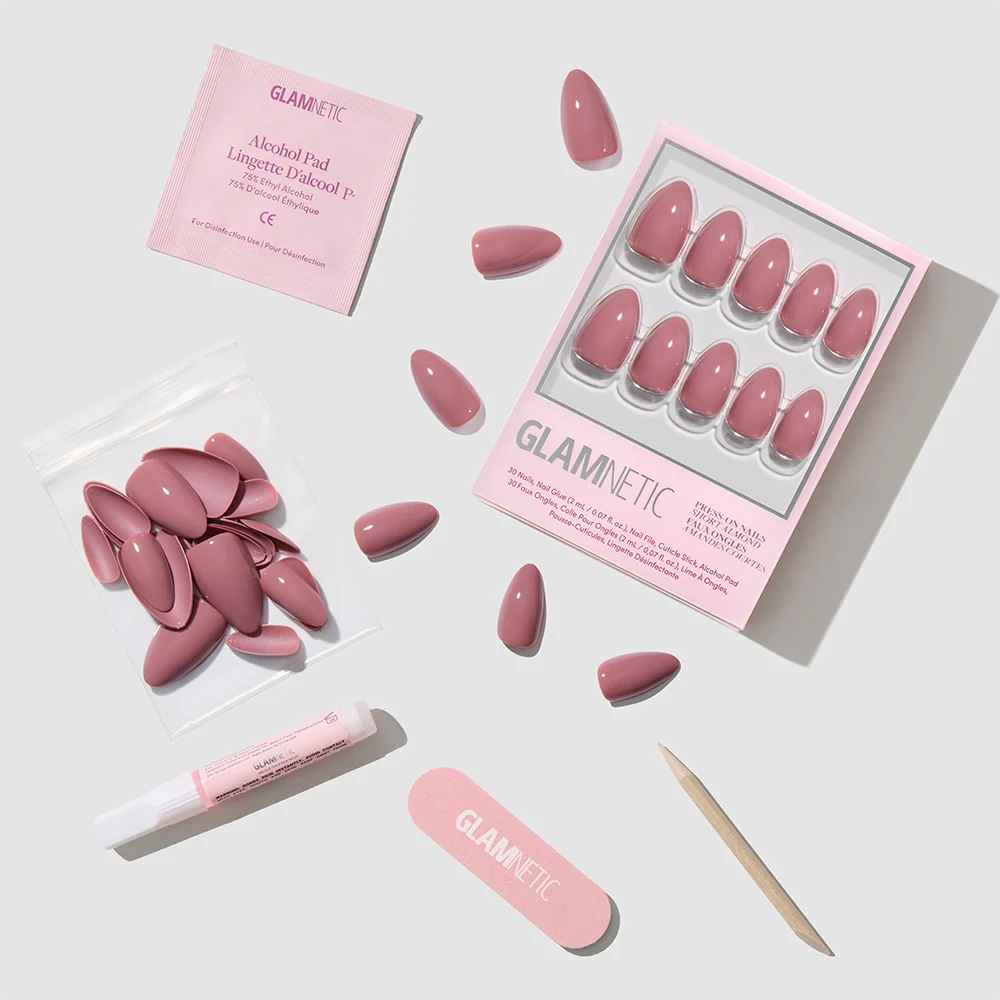 A flat lay of glamnetic's Mauve Obsession press-on nails in a glossy mauve finish, with nail glue, file, wooden cuticle stick, alcohol pad, and extra short almond nails arranged neatly on a light grey background.