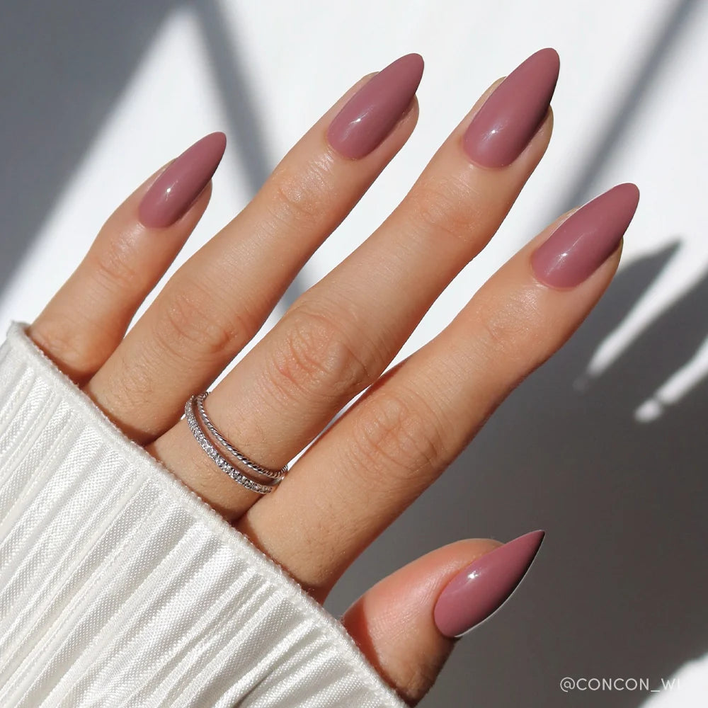 A hand with almond-shaped glamnetic Mauve Wife press-on nails, wearing stacked silver rings, rests on white ribbed fabric. Soft natural light and shadows add a minimal, elegant touch.