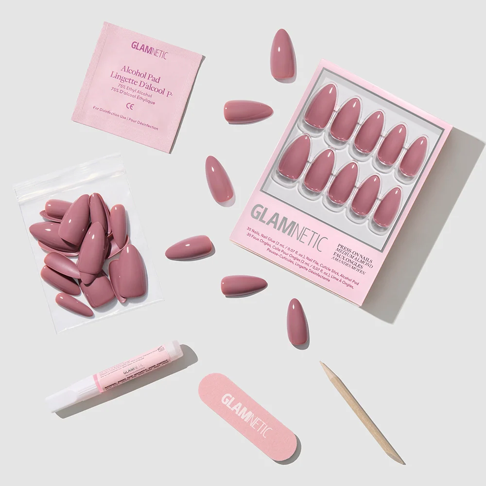 A set of Mauve Wife glossy pink fake nails, adhesive tube, wooden cuticle stick, pink glamnetic nail file, alcohol pad, and extra almond nails are arranged neatly on a light background. The packaging and file show the glamnetic brand.
