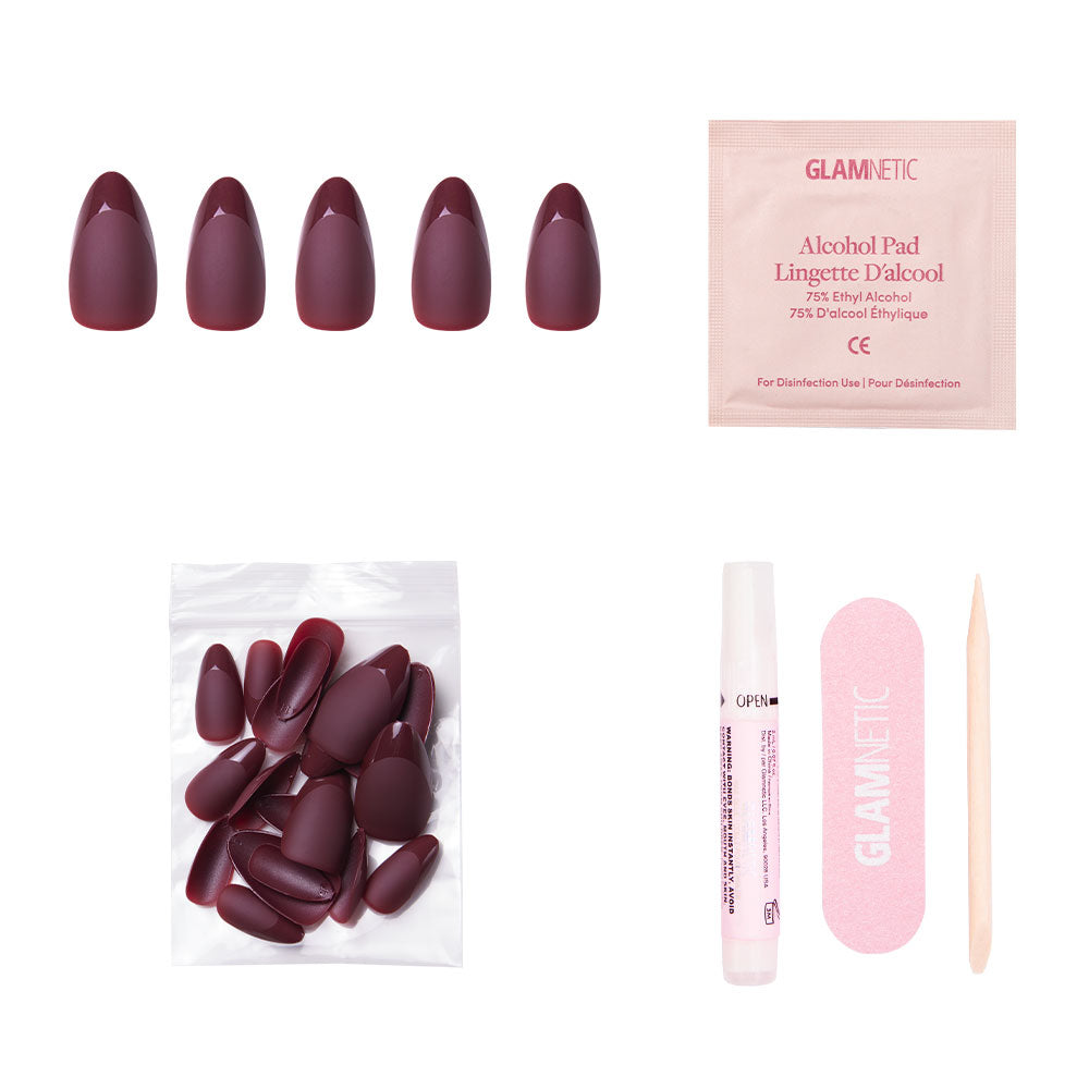 The Merlot nail set by glamnetic includes short almond-shaped burgundy matte press-on nails, extra nails, nail glue, an alcohol pad, a pink nail file, and a wooden cuticle stick—all arranged on a white background.