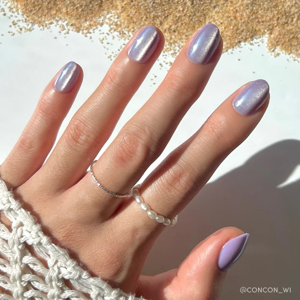 A hand wears glamnetic’s Mermaids Tail iridescent purple press-on nails, paired with silver and pearl rings, resting on white crochet fabric with golden beads scattered in the background.