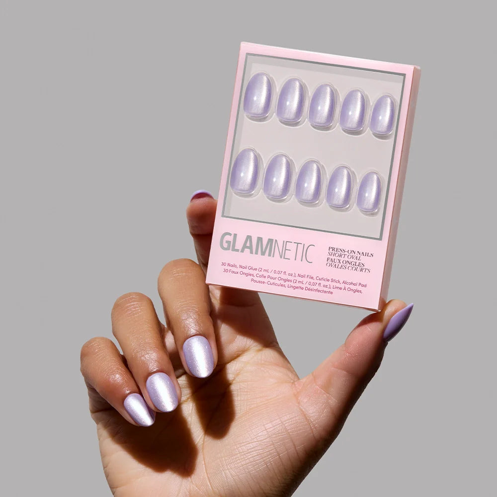 A hand with shimmery, iridescent purple Mermaids Tail press-on nails holds a glamnetic box featuring the matching set of nails against a plain grey background.