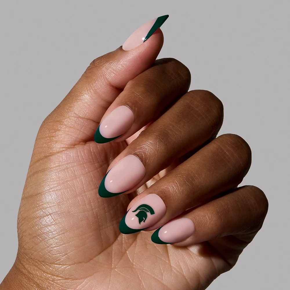 A hand with almond-shaped nails features the Michigan State Spartans Press-On Nail Set by glamnetic: a nude base, dark green French tips, and a Spartan helmet logo on the ring finger. The simple gray background highlights this spirited set.