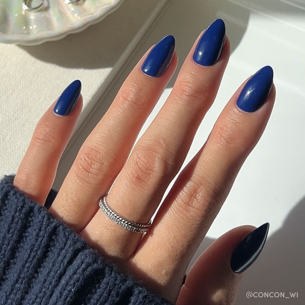A hand with long, almond-shaped nails in glamnetics glossy, vegan & cruelty-free Midnight Blue set. The person wears a dark sweater and silver double-band ring as sunlight highlights the nails.