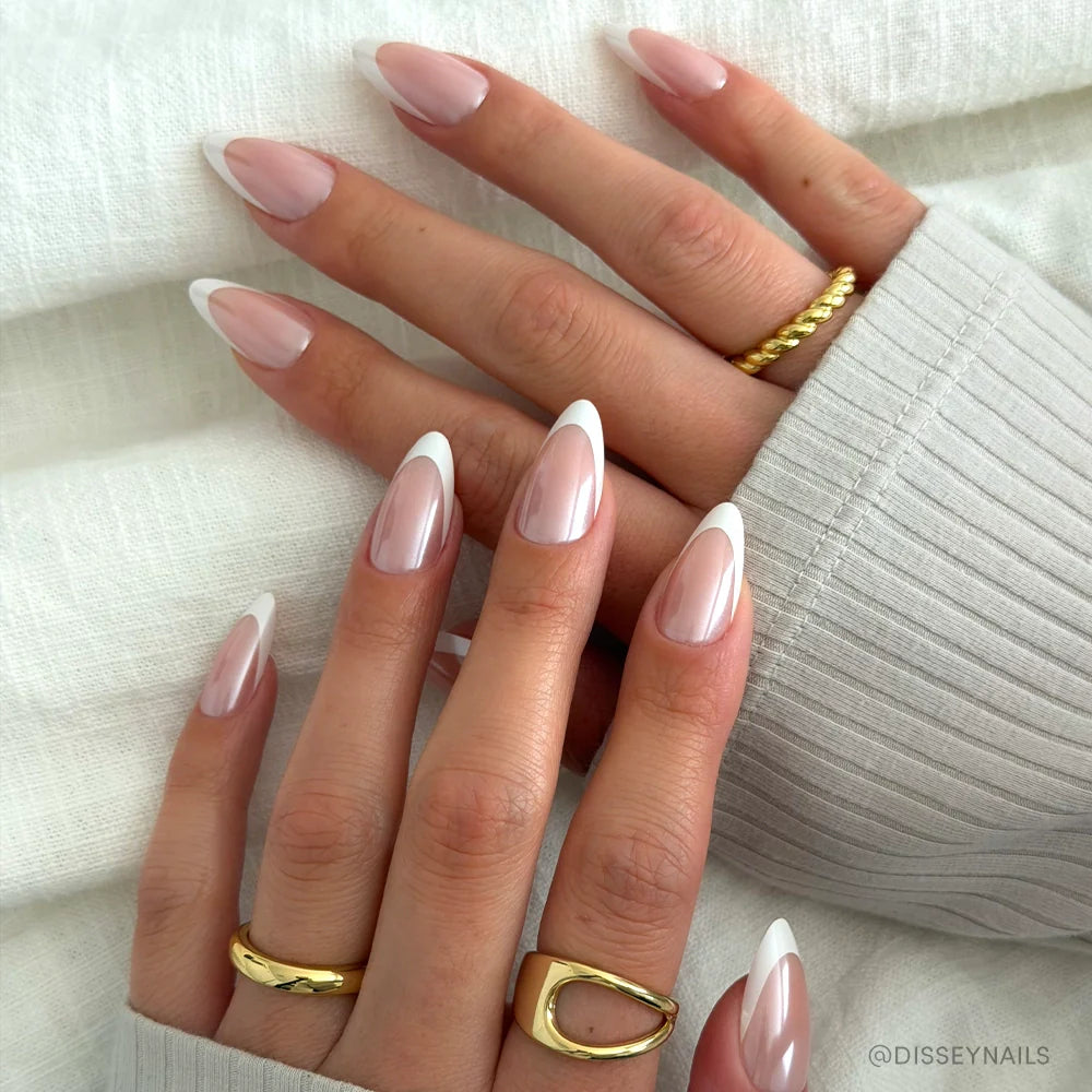 A close-up of hands with long, almond-shaped nails in glamnetic's Mother Pearl, showcasing a classic French tip and glossy finish. The hands wear gold rings and rest on textured white fabric.