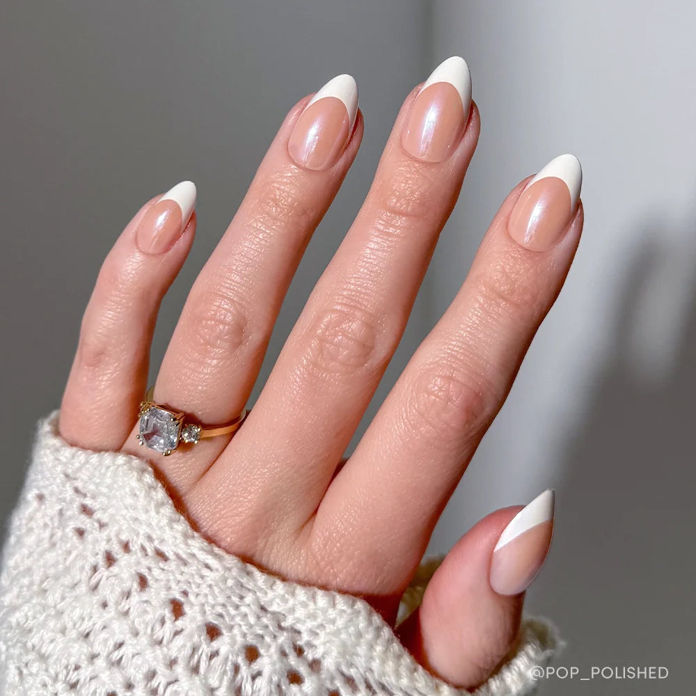 A hand with short almond nails sports glamnetic Mrs. press-ons in a modern French tip design, complemented by a gold gemstone ring and a textured white knit sleeve—perfect bridal nail inspo.