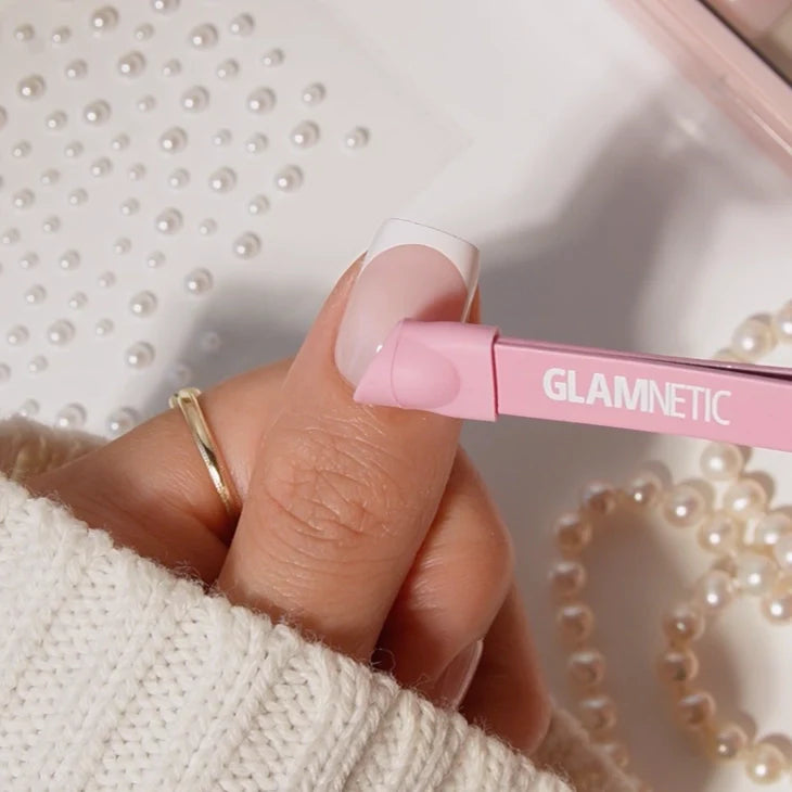 A hand with a gold ring and light pink French manicure uses glamnetic Nail Decal Tweezers to apply a pearl nail decoration, while pearl beads are scattered on a white textured surface in the background.