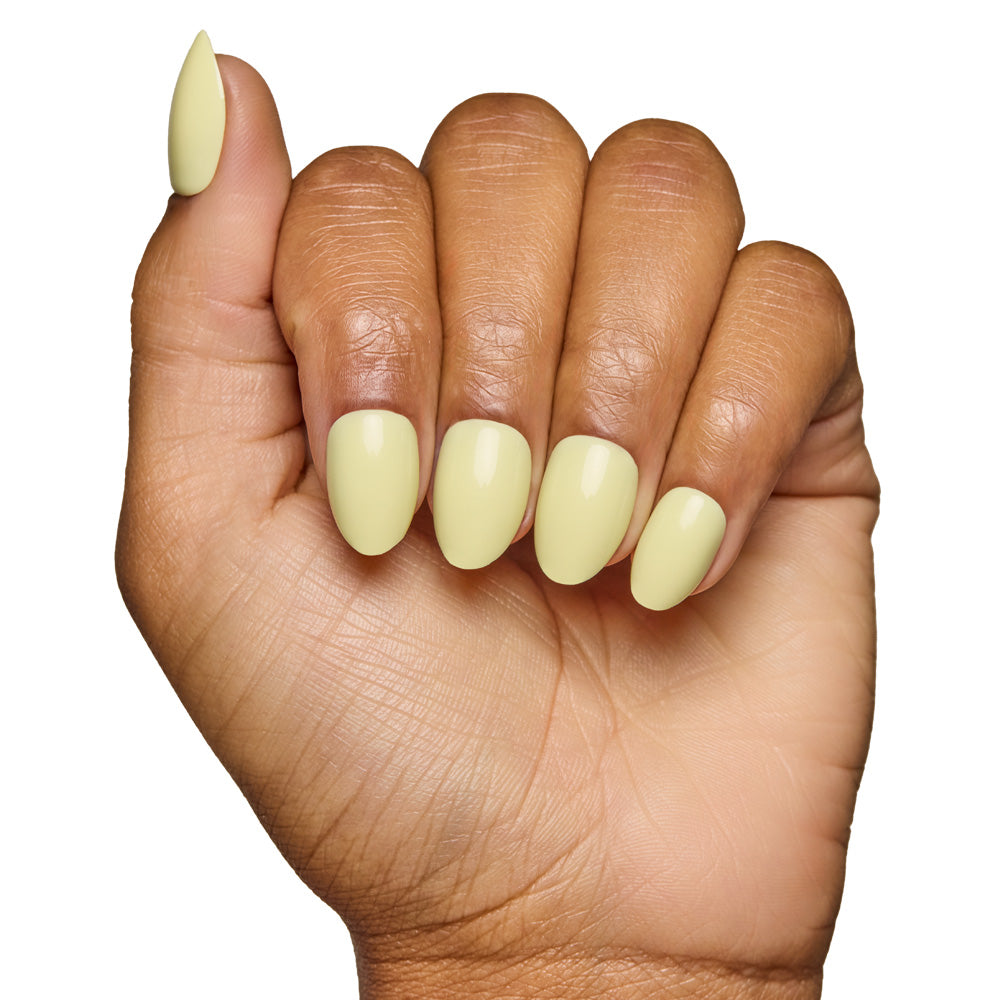 A hand with medium brown skin displays short oval nails in glamnetics Pastel Yellow, featuring a glossy, salon-quality finish. The fingers curl slightly against a white background, emphasizing the rounded almond shape.